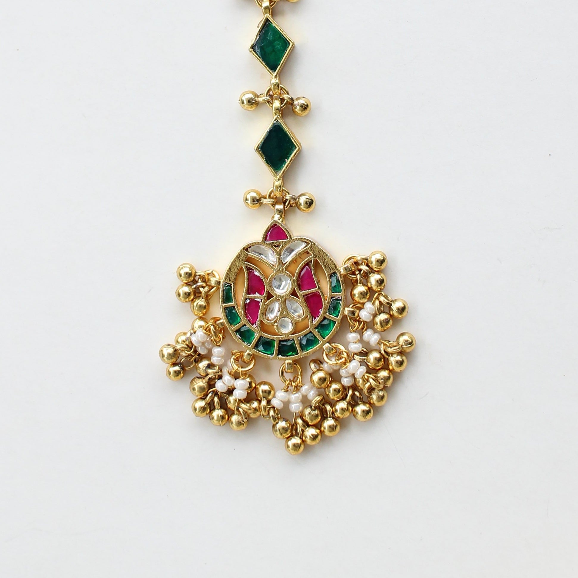 Gold-toned traditional Indian hair ornament (taali) with a detailed central motif featuring green and red stones, accented with small green diamond-shaped stones, displayed on a clean white surface.