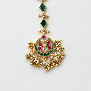 Gold-toned traditional Indian hair ornament (taali) with a detailed central motif featuring green and red stones, accented with small green diamond-shaped stones, displayed on a clean white surface.