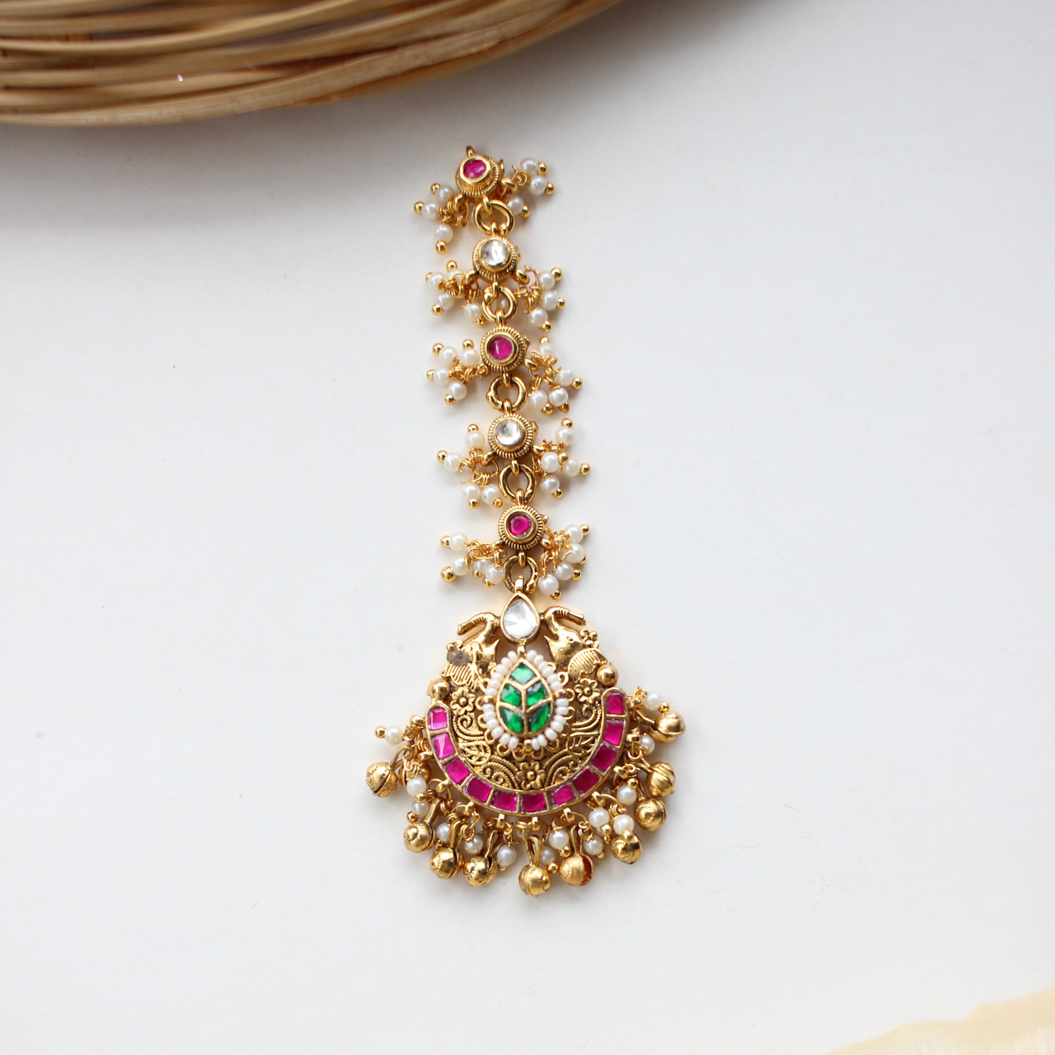Gold-toned traditional Indian jewelry piece featuring a central circular motif with a green and pink stone center, surrounded by gold beads and a row of matching small floral clusters, displayed on a white surface with decorative fabric and wicker props.
